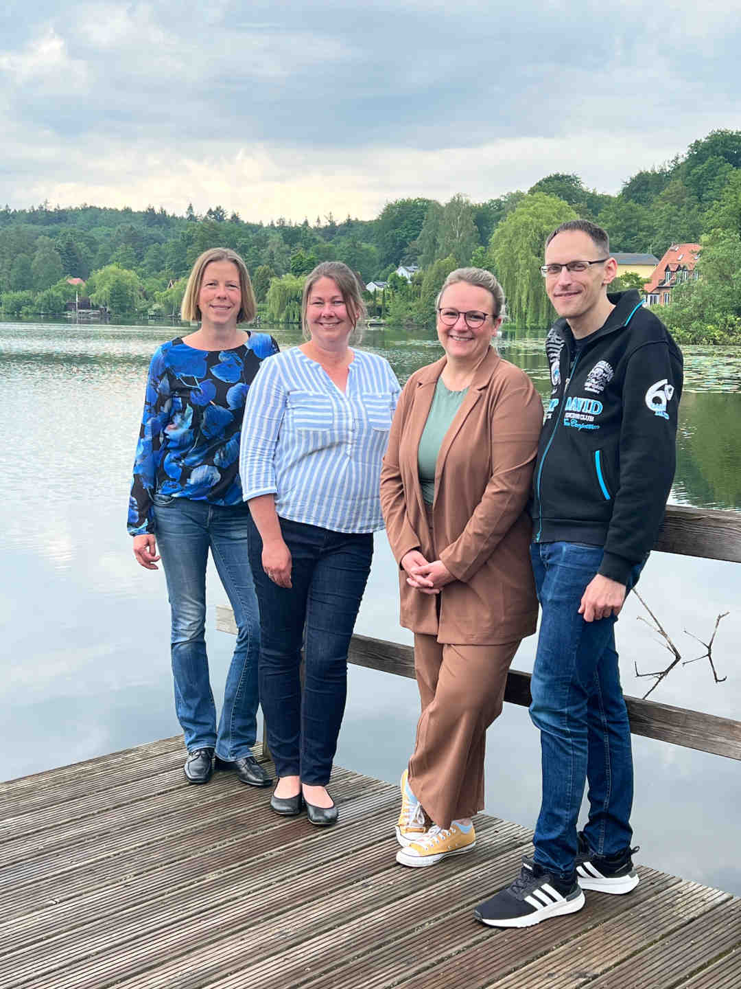 Das Team vom Koordinationsbüro Mölln Inklusiv, bestehend aus vier Menschen, steht nebeneinander auf einem Steg an einem See. Im Hintergrund sind Wälder und Dächer von Häusern zu sehen und der Himmel ist leicht bewölkt.