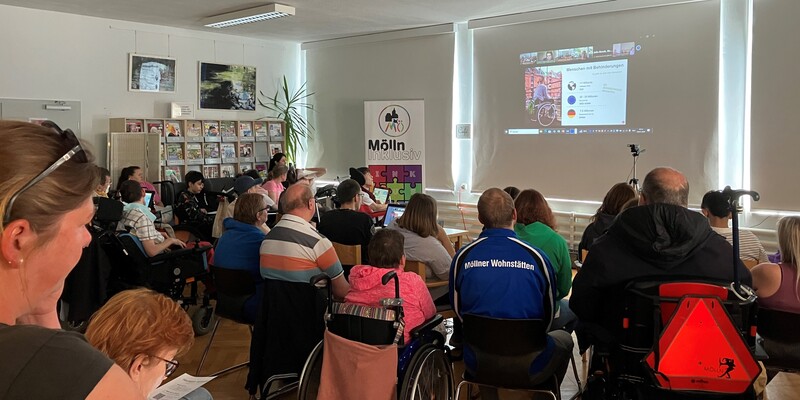 Eine Menschengruppe sitzt in einem Halbkreis. Sie nehmen an einer Veranstaltung teil. Ein Beamer zeigt ein Bild an der Wand. Die Menschengruppe schaut aufmerksam auf das Bild.