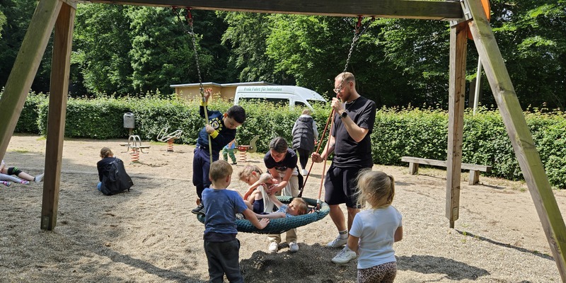 Kinder spielen auf einem Spielplatz. Zwei Kinder liegen gemeinsam in einer Nestschaukel. Es sind Erzieher*innen dabei.