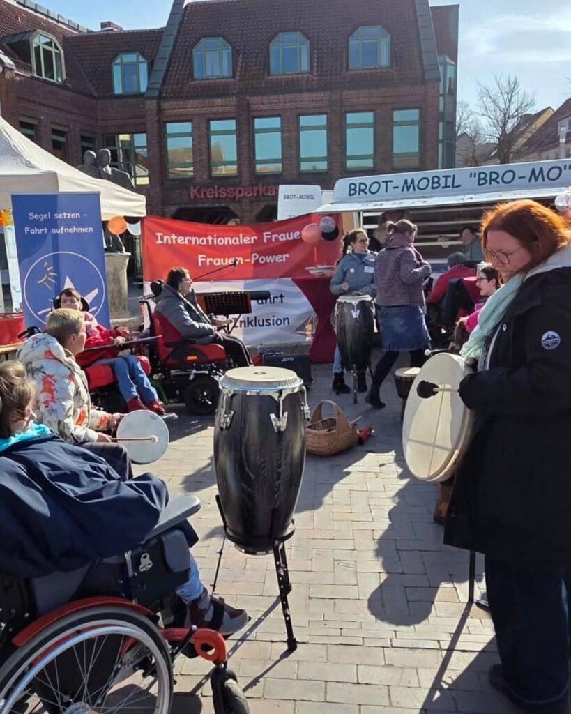 Eine Gruppe von Menschen, darunter Rollstuhlfahrer, steht auf einem Event mit Trommeln und Musikinstrumenten. Im Hintergrund ist ein Banner mit der Aufschrift 'Internationaler Frauentag - Frauen-Power' und Hinweise auf Inklusion. Es herrscht eine festliche Atmosphäre.