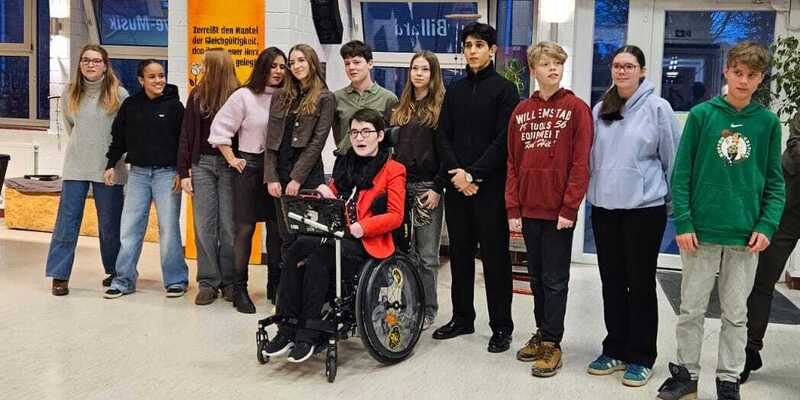 Gruppe von Jugendlichen und jungen Erwachsenen in einem Innenraum, einige stehen, eine Person sitzt im Rollstuhl, im Hintergrund bunte Plakate an der Wand