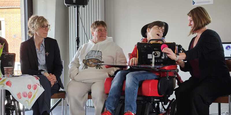 Drei sitzende Personen, darunter ein Dame im Rollstuhl mit einem Bildschirm vor sich, eine Frau rechts bedient den Bildschirm, links ein Tisch mit einer bunten Tischdecke