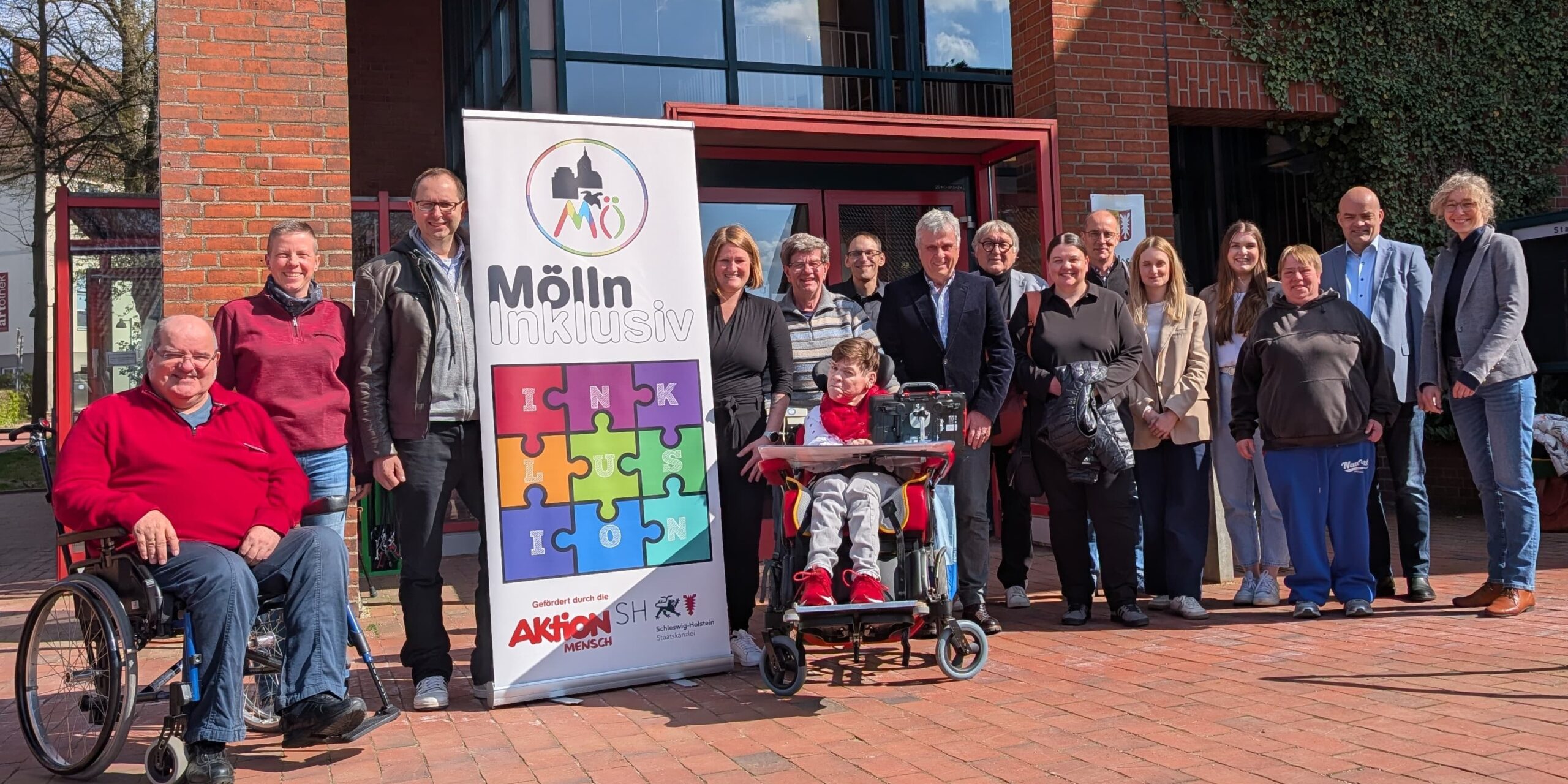 Gruppe von Menschen, darunter zwei Personen im Rollstuhl, steht vor einem Gebäude neben einem Banner mit buntem Puzzle und der Aufschrift 'Mölln inklusiv'
