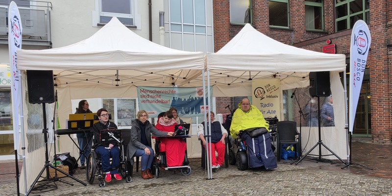 Menschen mit und ohne Rollstuhl sitzen unter zwei weißen Pavillons vor einem Backsteingebäude mit Bannern und Lautsprechern