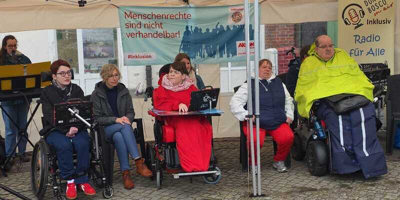 Gruppe von Menschen unter einem Pavillon, darunter drei Personen im Rollstuhl, mit Bannern im Hintergrund zu Menschenrechten und Inklusion.