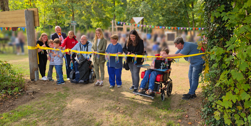 Gruppe von Erwachsenen und Kindern, darunter zwei Rollstuhlfahrer, schneidet gemeinsam ein gelbes Band in einem Park mit Bäumen und Wimpelgirlanden durch